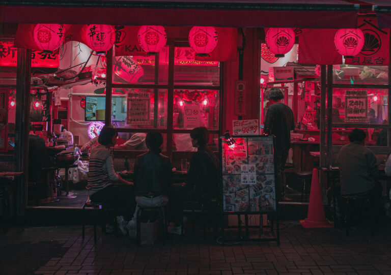 night view of a cozy tokyo restaurant, glowing red lanterns hang above as locals sit inside, enjoying food and conversation